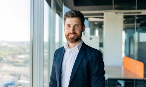 Successful person. Handsome businessman in formal wear standing in office near window, posing looking and smiling at camera, free space